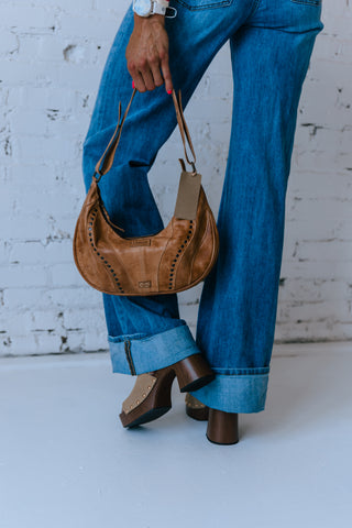 Person wearing blue jeans and brown shoes holding a brown handbag against a white brick wall.