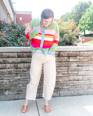 A woman stands outdoors in front of a brick wall and flowering bushes, wearing a pastel striped knit cardigan with cream pinstriped cropped pants and tan wedge sandals.