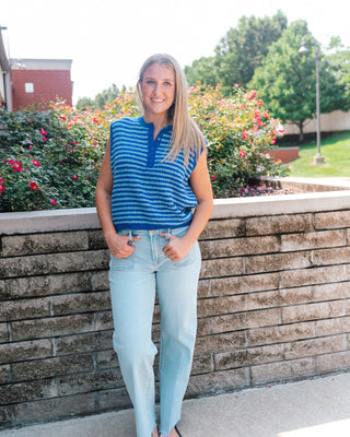 A woman stands outdoors in front of a stone wall and flowering bushes, wearing a blue and white striped sleeveless top with a polo collar and light-wash jeans.