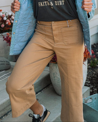 A woman stands on a front porch wearing a black graphic T-shirt with a bull skull and the words 'LIVING ON FAITS & DIRT,' a light blue quilted jacket, and tan wide-leg pants. An American flag is visible in the background.