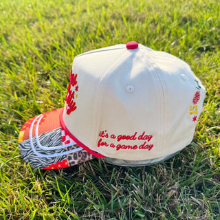 A white baseball cap with red embroidered text reading 'it's a good day for a game day'. The cap features a red button on top, red embroidery, and a patterned brim with red, white, black, and leopard print.