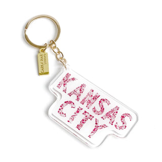 A clear acrylic keychain featuring the words 'KANSAS CITY' in a pink FLORAL font, attached to a gold-tone split ring and small hanging tag 